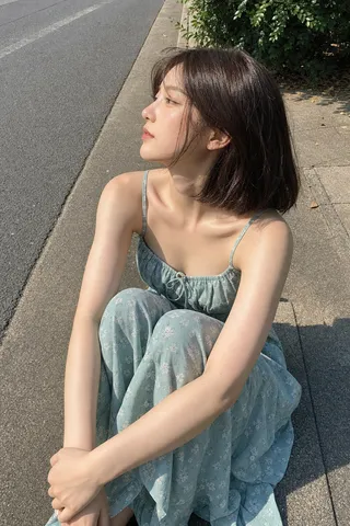 Profile shot of a young woman with short brown hair sitting on a street curb hugging her knees in sunlight.