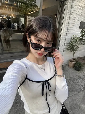 Young woman with bob haircut pulling down sunglasses for a selfie outside a café.