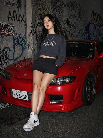 Woman in dark sweatshirt and shorts leaning against a red sports car under direct flash.