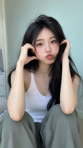 Close-up portrait of a woman in a white ribbed tank top posing playfully by a light aqua wall.