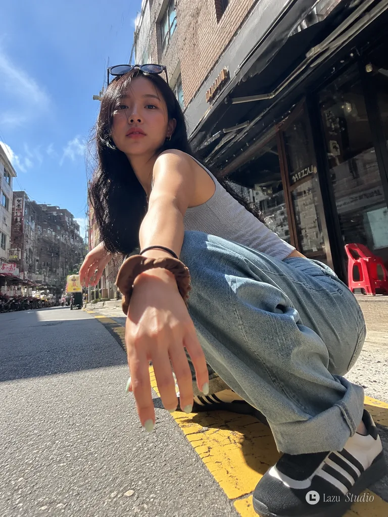 Dynamic low-angle street photograph of a stylish young woman squatting on an urban sidewalk in the sun.