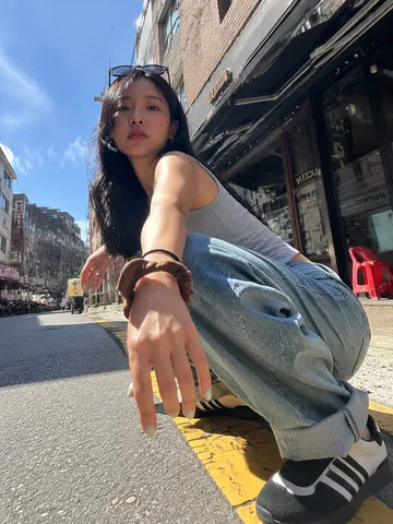 Dynamic low-angle street photograph of a stylish young woman squatting on an urban sidewalk in the sun.
