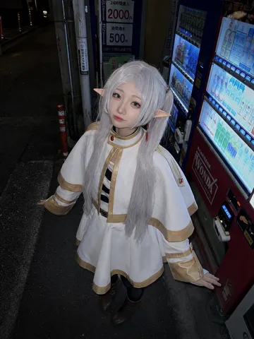 Woman in Frieren cosplay looking up at the camera beside illuminated vending machines at night.