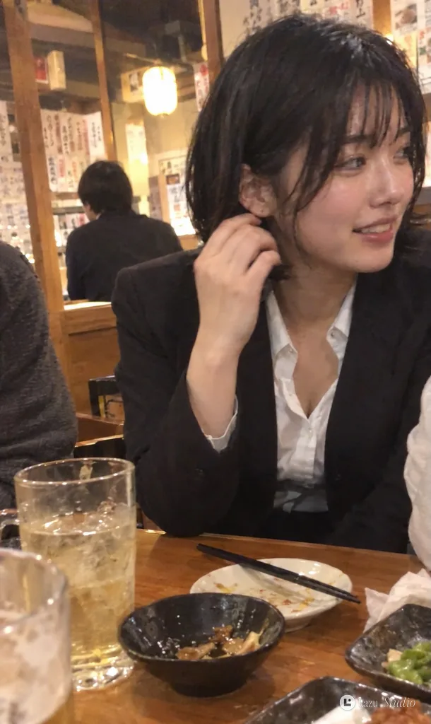 Candid smartphone screenshot of a young woman smiling loosely at a warm izakaya table.