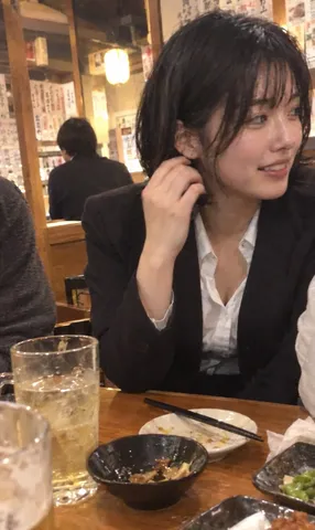 Candid smartphone screenshot of a young woman smiling loosely at a warm izakaya table.
