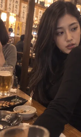 A casual, grainy vertical photo of a young woman seated at a wooden izakaya table with dishes.