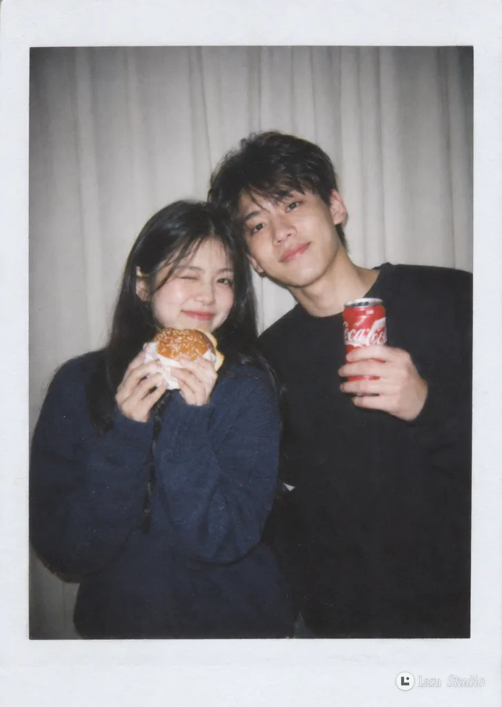 A smiling young couple posing side by side under direct flash, holding a burger and a can of soda.