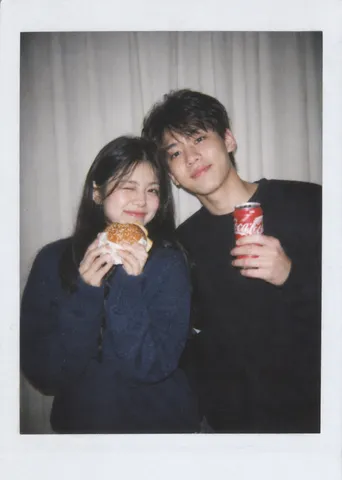 A smiling young couple posing side by side under direct flash, holding a burger and a can of soda.