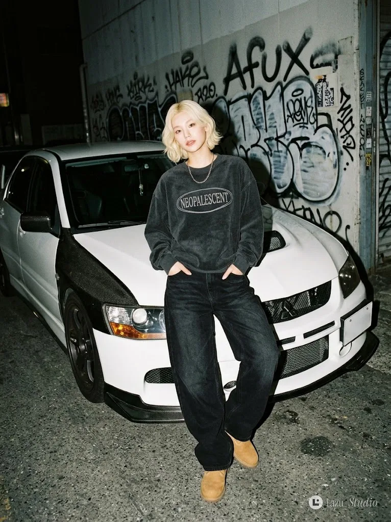 A woman in an oversized sweatshirt leaning on a white sports car against a graffiti wall at night.