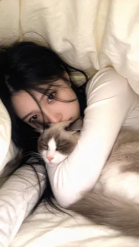 Close-up lo-fi photo of a young woman lying under a blanket holding a cat, with only her eyes visible.