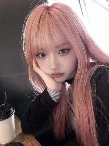 Close-up selfie of a young woman with pastel pink hair leaning her cheek on her hand in a café.