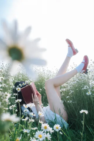 Woman in a white dress lying in a blooming daisy meadow with a book over her face.