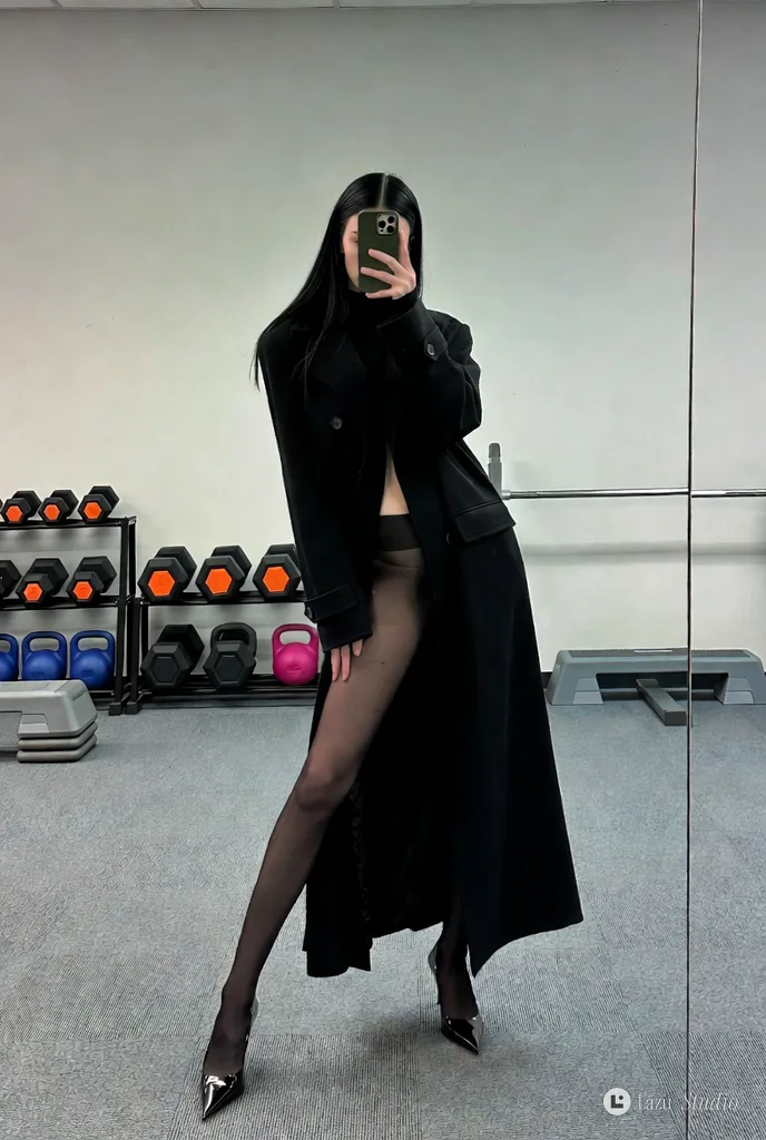 Full-length mirror selfie of a woman in a black coat and heels with her face obscured by a phone.