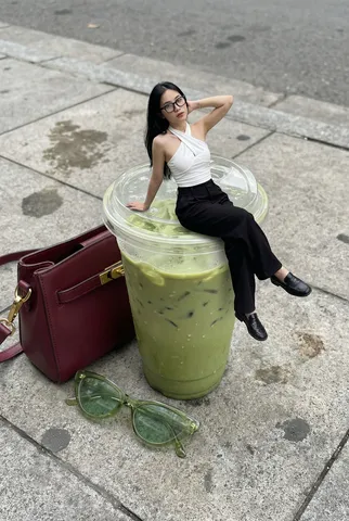 A miniature woman sitting relaxed on the rim of a clear plastic cup filled with iced green matcha.