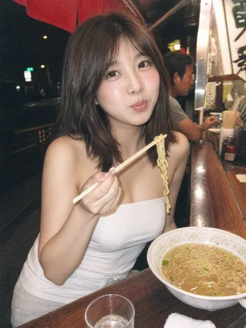 High-angle direct flash photo of a woman holding chopsticks with noodles at an outdoor food stall.