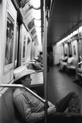 Black and white photo of a cowboy sleeping on an NYC subway car seat with his hat over his face.
