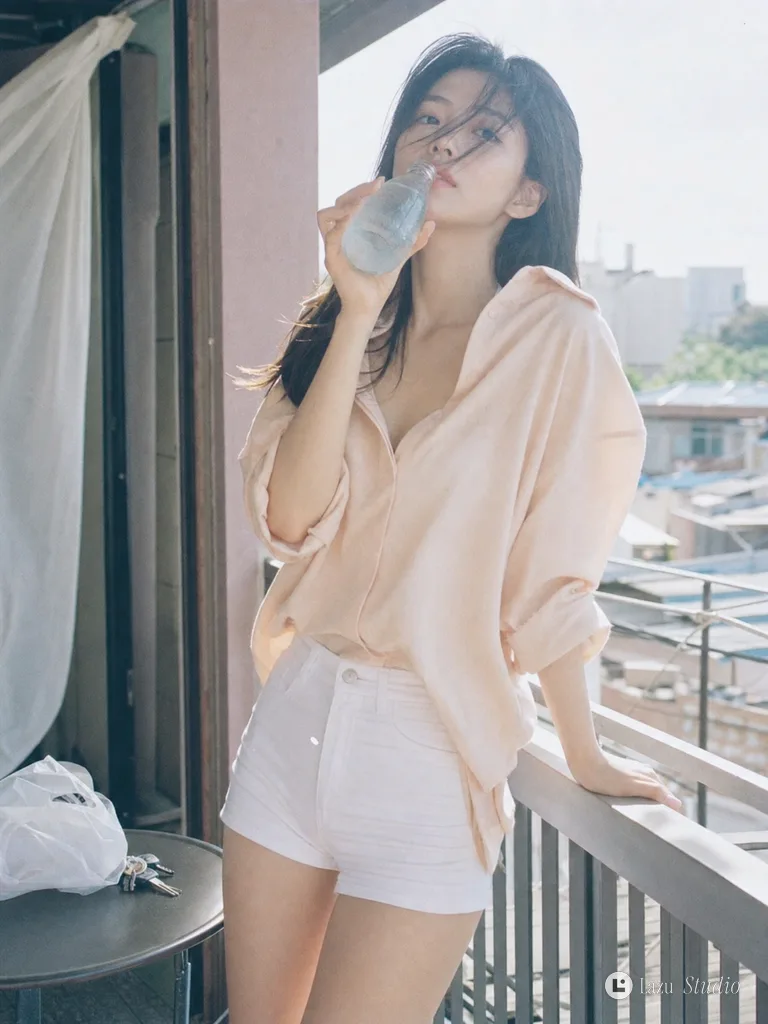 Young woman in an oversized peach shirt leaning against a summer balcony railing holding a water bottle.