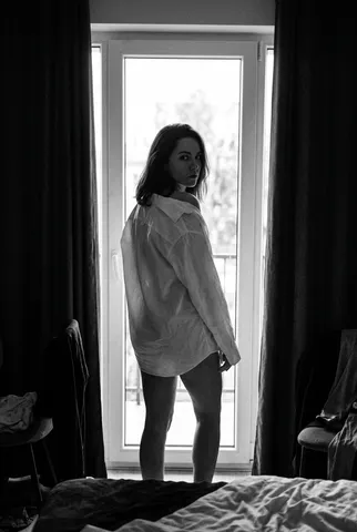 Monochrome silhouette of a woman in an oversized shirt standing before a bright window and looking back.