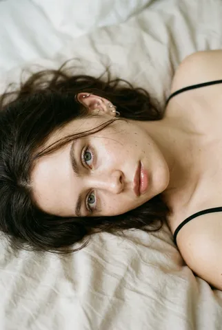 Close-up portrait of a young freckled woman lying on light bedding, looking softly at the camera.