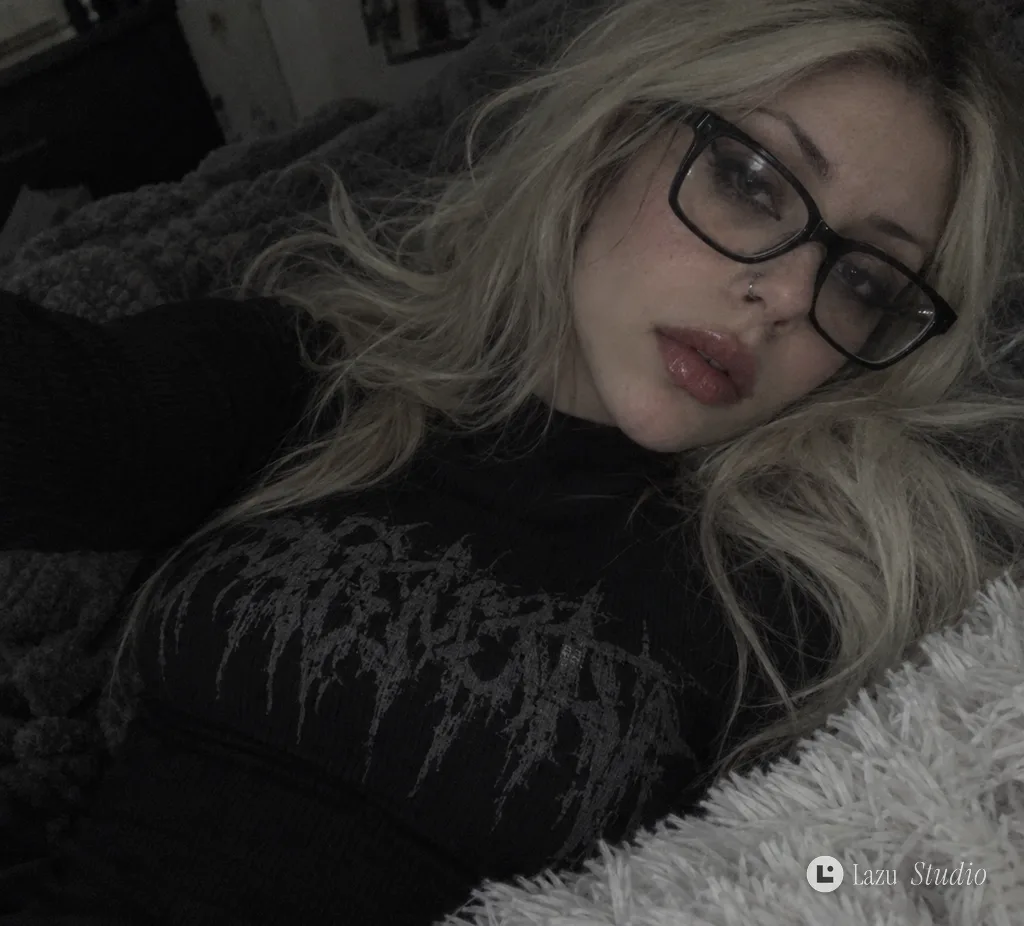 Tilted selfie of a woman wearing glasses and a dark gothic top lying on a textured blanket.
