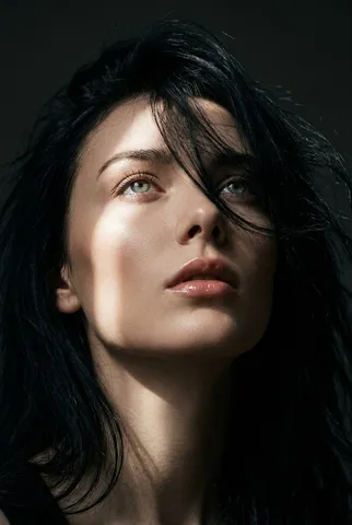 Close-up portrait of a woman with windswept hair across her face, highlighted by a diagonal light beam.