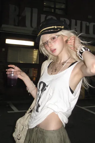 Young woman with platinum hair and black cap holding a purple drink outside a brick storefront at night.