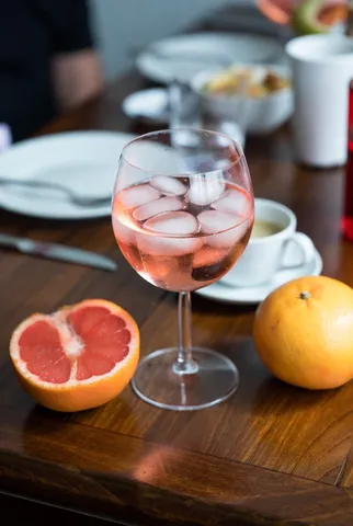 A glass of pink gin with ice and grapefruit slices sitting on a casual, cluttered wooden table in daylight.